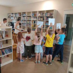 dzieci stoją wsród bibliotecznych regałów i słuchają lekcji bibliotecznej